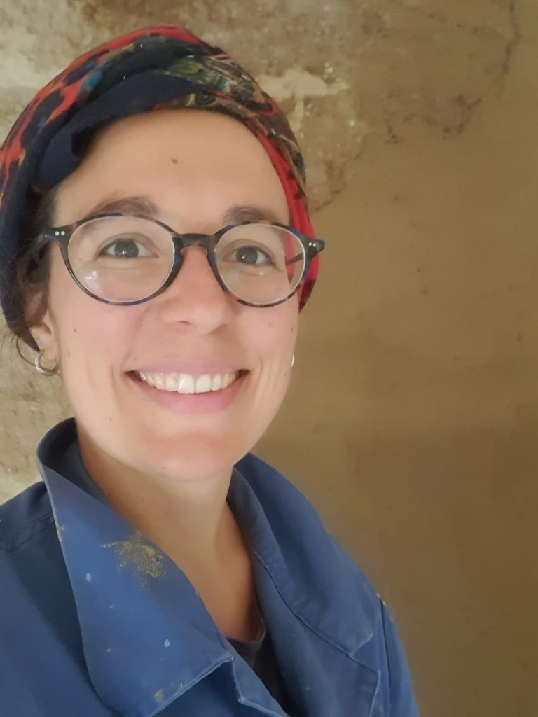 Portrait souriant de Cécile, artisane enduiseuse, devant un mur en pisé en train d'être enduit en terre